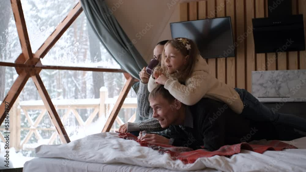 Family of four enjoying winter vacation in dome camping, relaxing on bed over big window with snowy winter nature