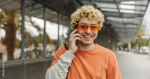 Smiling blonde young caucasian guy in orange sunglasses talking on phone. Curly male with panoramic windows in background. Concept emotions, gadgets, lifestyle.
