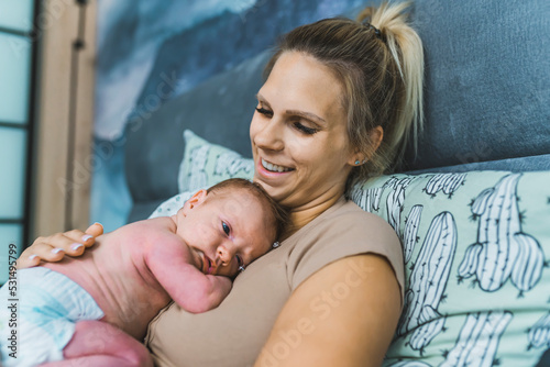 Infant baby in diaper slowly falling asleep while lying down on his mother's chest. Happy caucasian blond mother smiling at baby. Bedroom interior. High quality photo