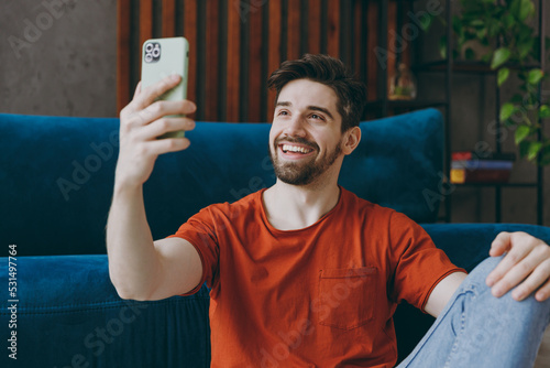 Young man wear red t-shirt do selfie shot on mobile cell phone post photo on social network sit on blue sofa coch stay at home flat rest relax spend free spare time in living room indoors grey wall. photo