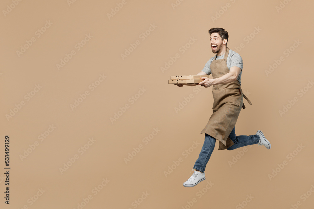 Full size young man barista barman employee wear brown apron work in coffee shop jump run hold pizza in cardboard flatbox isolated on plain pastel light beige background Small business startup concept