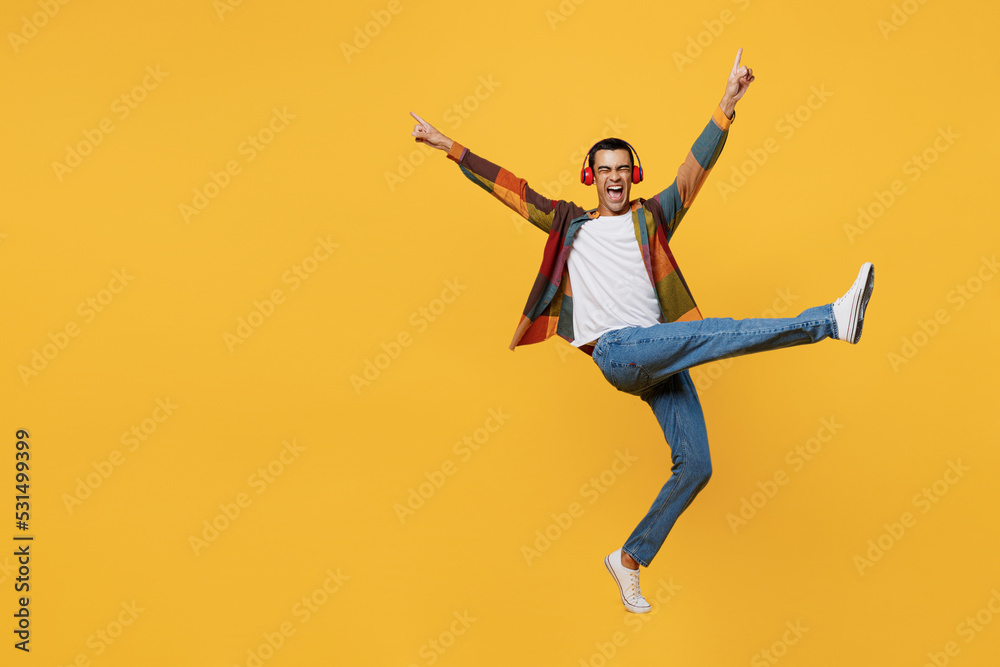 Full body young middle eastern man 20s he wear casual shirt white t-shirt headphones listen music dance raise up hands leg isolated on plain yellow background studio portrait People lifestyle concept.