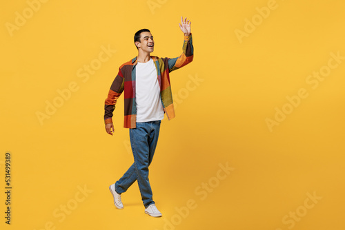 Full body side view young smiling happy middle eastern man 20s wear casual shirt white t-shirt walking going strolling waving hand isolated on plain yellow background studio People lifestyle concept.