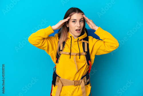 Young mountaineer woman with a big backpack isolated on blue background with surprise expression