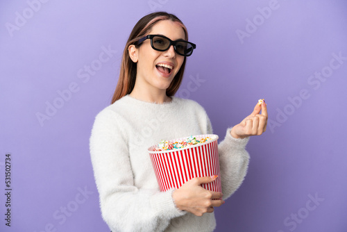 Young caucasian woman isolated on blue background with 3d glasses and holding a big bucket of popcorns