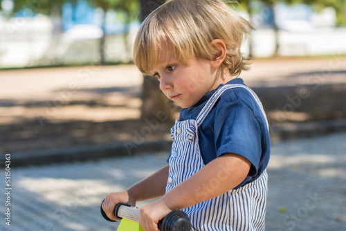 Portrait of an adorable 2 year old blond boy smiling while playing in the park with a tricycle. Background out of focus. Side view of a toddler playing