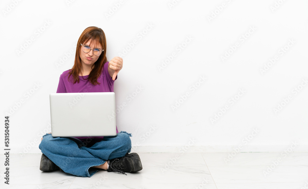 redhead girl with a laptop sitting on the floor showing thumb down with negative expression redhead girl with a laptop sitting on the floor showing thumb down with negative expression