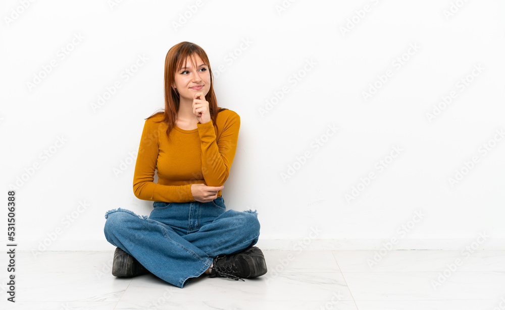 Redhead girl sitting on the floor isolated on white background looking to the side Redhead girl sitting on the floor isolated on white background looking to the side