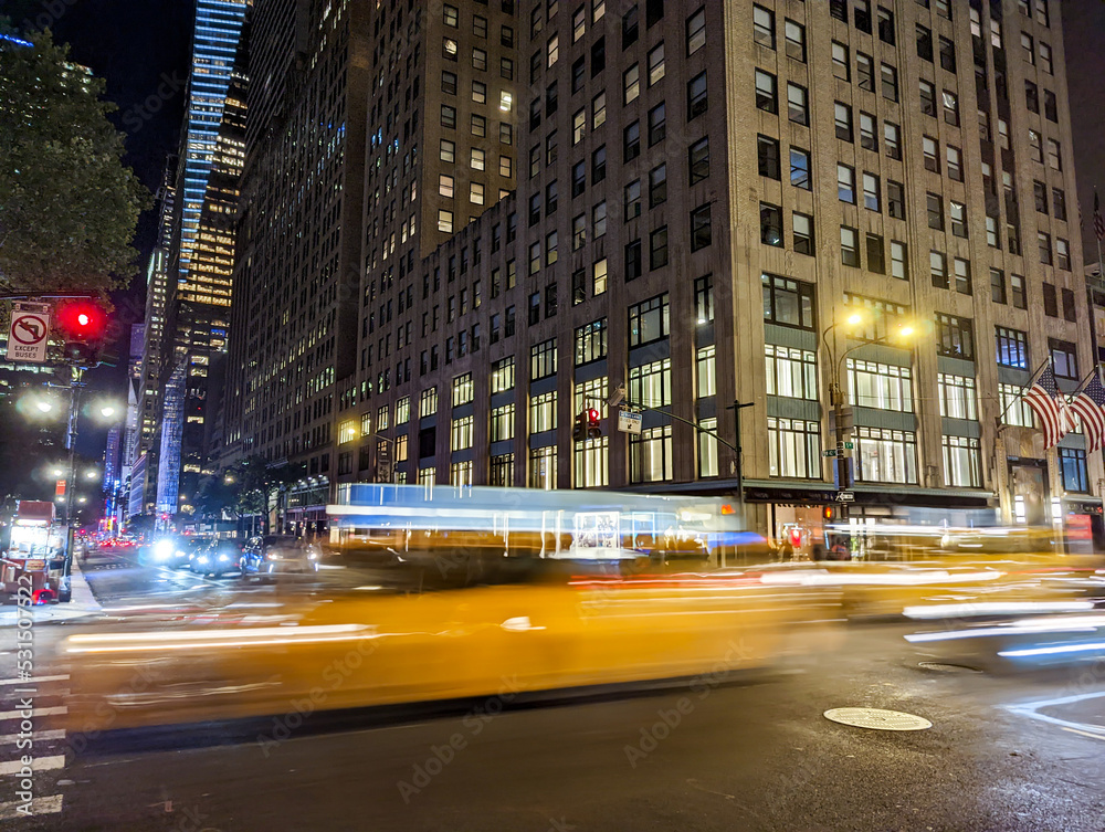 Blurred motion of a yellow taxis driving down the street through New York City at night with building lights in the background