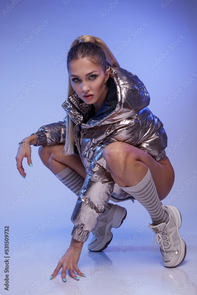 Fototapeta premium High fashion photo of a beautiful elegant young woman in a pretty blue bodysuit, silver metallic coat jacket, high socks, sneakers on blue background. Studio Shot.