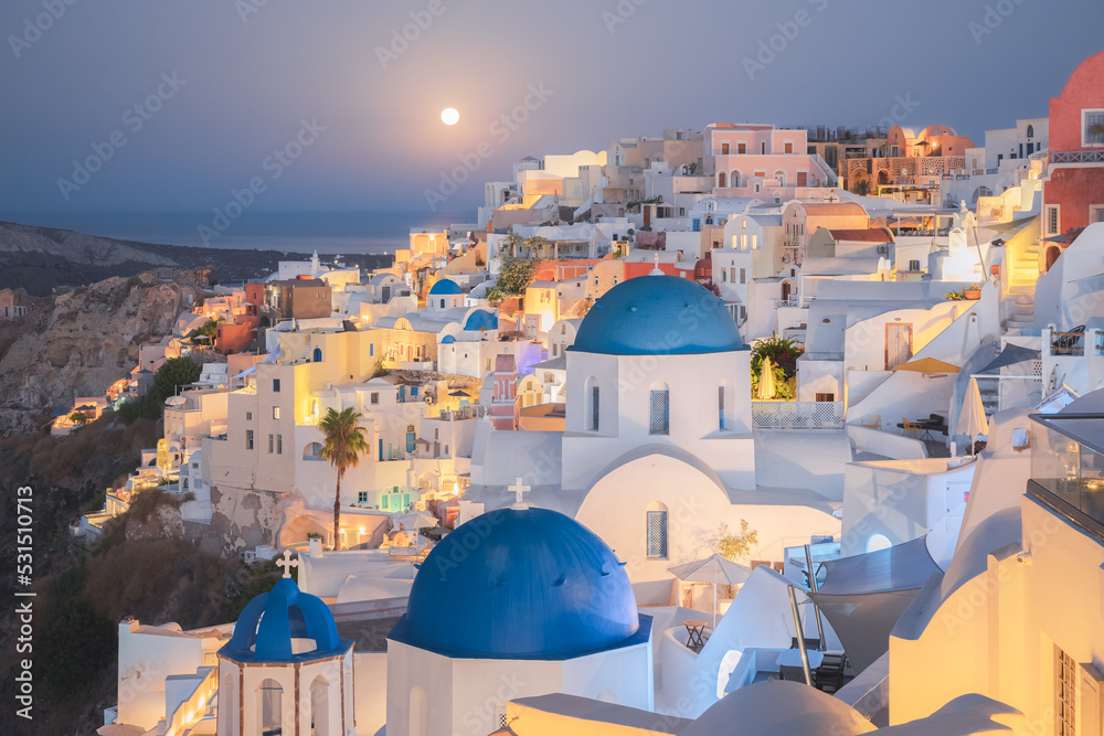 Seaside moonlit view at night of traditional white wash buildings and blue dome churches at the popular seaside tourist resort village of Oia on the Greek island of Santorini, Greece.