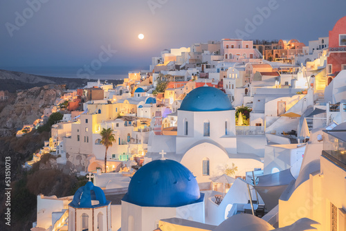 Seaside moonlit view at night of traditional white wash buildings and blue dome churches at the popular seaside tourist resort village of Oia on the Greek island of Santorini, Greece.
