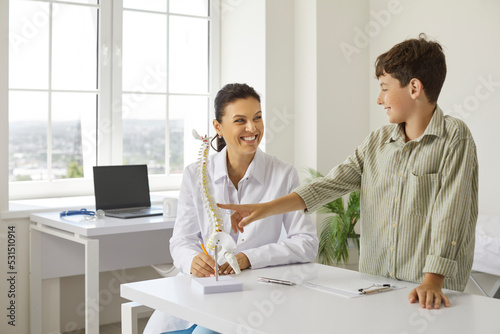 Happy, friendly, smiling doctor at orthopedic clinic communicating with child, showing anatomical spine model, telling about skeleton and spinal curvatures and explaining how to prevent back problems