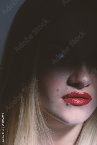 Fashion and make-up concept. Beautiful blonde woman portrait partly face covered with shadow. Mouth and nose illuminated with light. Looking at camera. Red lipstick make-up. Toned image in beige color