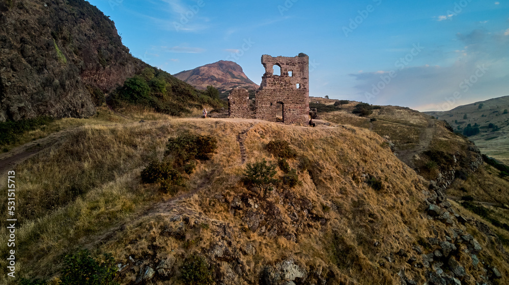 Ruin of St Anthony Chapel in summer evening with top of Arthur Seat at distance - drone photo