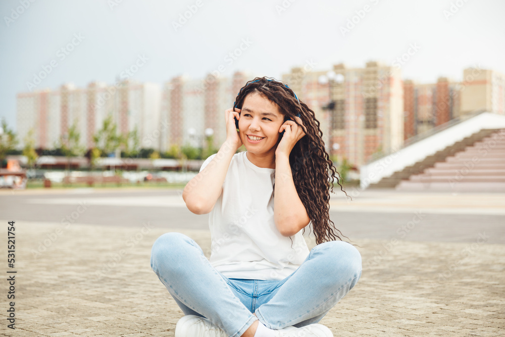 A smiling girl with dreadlocks in headphones is sitting on the floor in the park. A happy young woman relaxing with headphones, enjoying music. Space for copying.generation z A smiling girl with dreadlocks in headphones is sitting on the floor in the park. A happy young woman relaxing with headphones, enjoying music. Space for copying.generation z