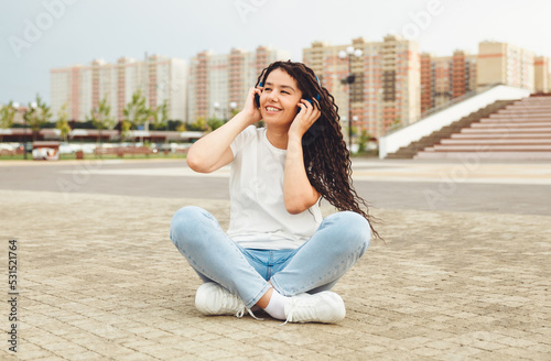 A smiling girl with dreadlocks in headphones is sitting on the floor in the park. A happy young woman relaxing with headphones, enjoying music. Space for copying.generation z © inna717