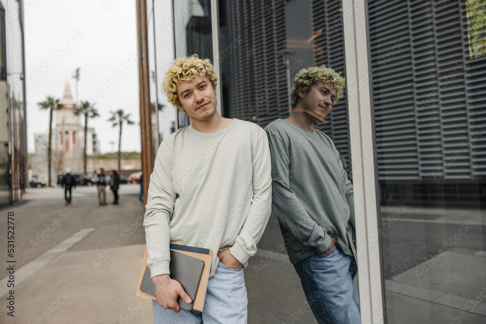 Young caucasian guy standing near glass wall with folder and tablet. Curly man dressed in casual clothes outdoor. Concept fashion, gadgets.