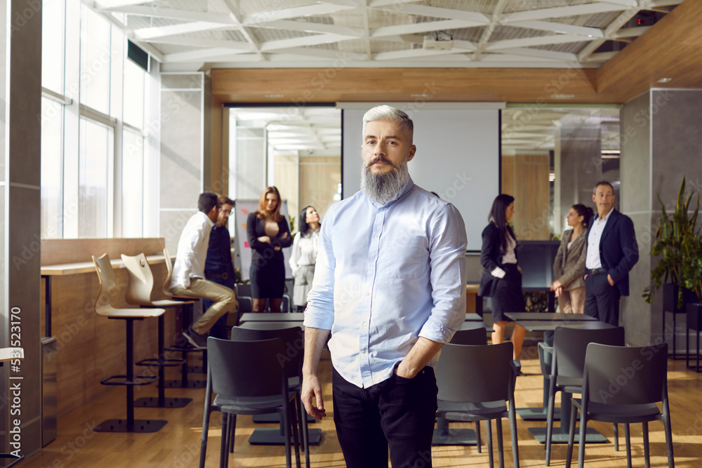 Portrait of serious business teacher and professional team coach. Handsome man with white hair and grey beard standing in office after corporate training class, with group of employees in background