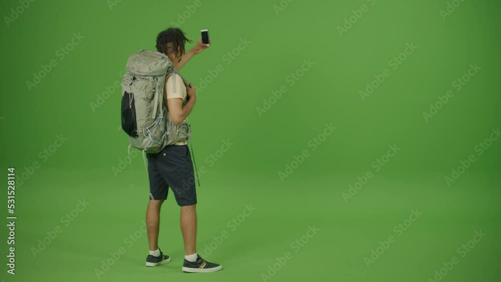 Happy Mexican Curly Tourist with Big Travel Backpack Looking to The Left, Making Photo and Video around Him on His Smartphone, Making Selfie and Smiling on Green Screen Background Happy Mexican Curly Tourist with Big Travel Backpack Looking to The Left, Making Photo and Video around Him on His Smartphone, Making Selfie and Smiling on Green Screen Background