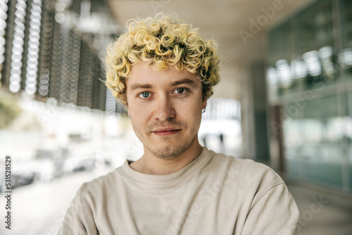 Close-up of calm young caucasian guy looking at camera with serious face on street. Curly blonde takes part in portrait shoot. Conceptual photography.