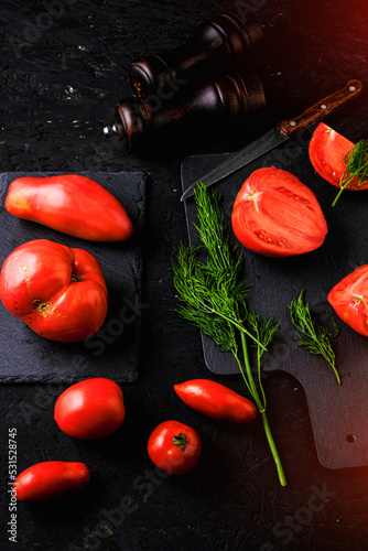 Sliced tomatoes on a slate, dill greens, two spice mills and knife with wooden handle on dark background.