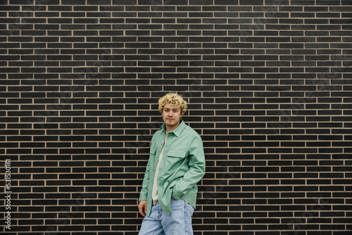 Calm young caucasian guy dressed green shirt keeping his hand in pocket. Curly man standing near brick wall. Concept fashion, lifestyle. © Look!