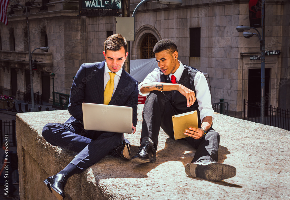 Two Young Men with laptop computer and tablet computer working, reading together on Wall Street in New York City..