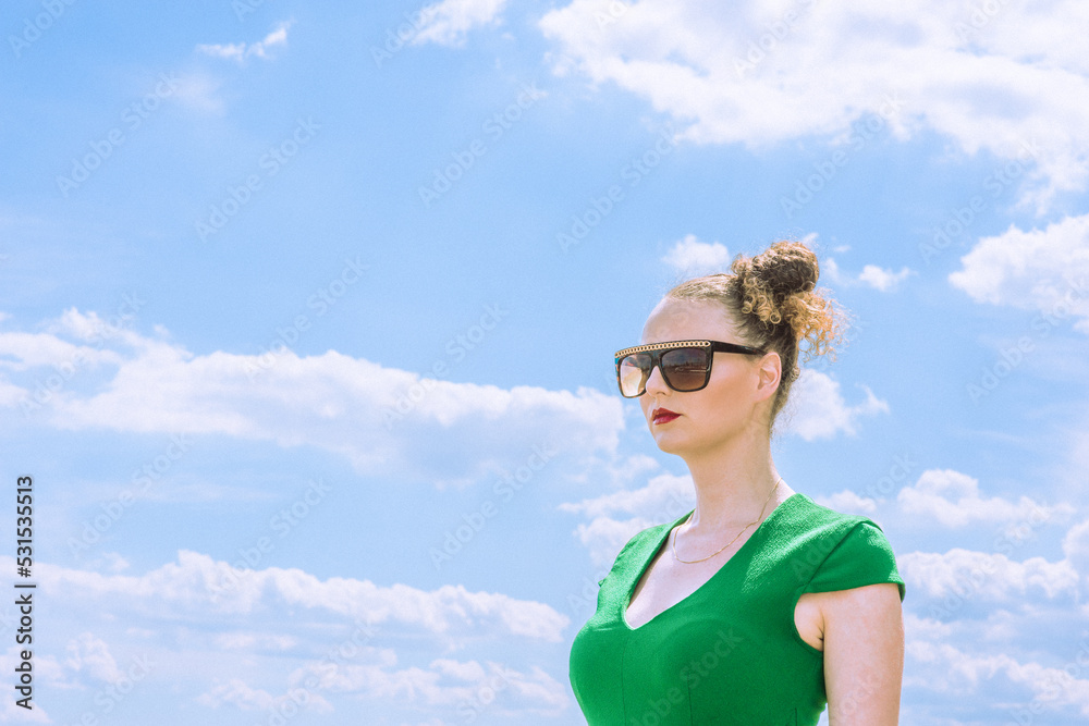 Young American woman, wearing sunglasses, dressing in green, standing under sunny sky in hot summer..