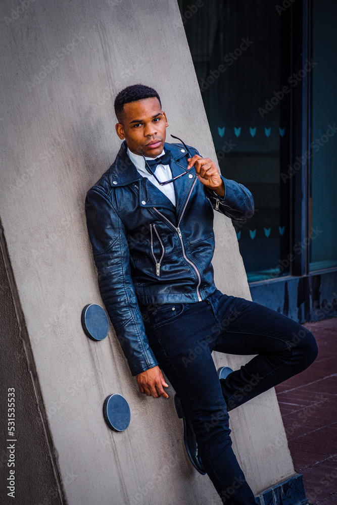 Young African American Man, wearing black leather jacket, jeans, white undershirt, bow tie, holding sunglasses, standing against wall on street in New York City..