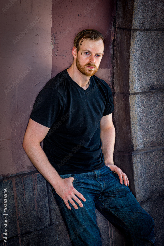 Young man with beard, wearing black v neck T shirt, jeans, standing against painted wall outdoors in New York City, relaxing, .