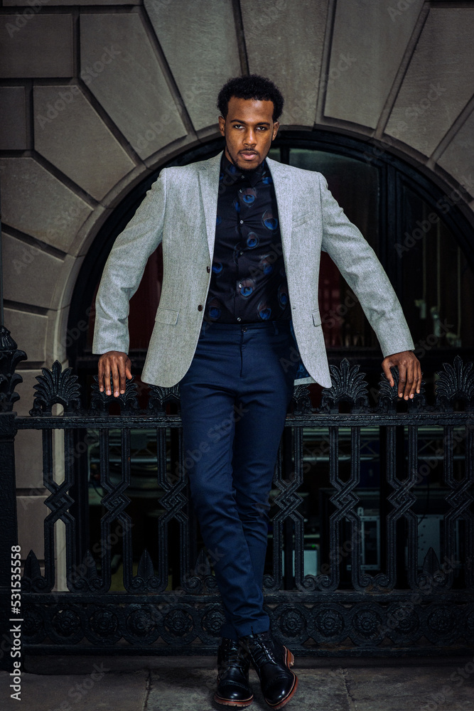 Young African American man wearing gray blazer, flower patterned undershirt, black pants, leather boot shoes, standing by railing outside office building in New York City, looking forward..