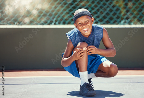 Child with a band aid on a knee injury from sports outside on a basketball court touching his bandage. Boy with a medical plaster hurt by accident while training or practicing for a game.