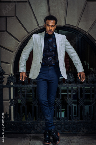 Obraz na płótnie Young African American man wearing gray blazer, flower patterned undershirt, black pants, leather boot shoes, standing by railing outside office building in New York City, looking forward.. w salonie