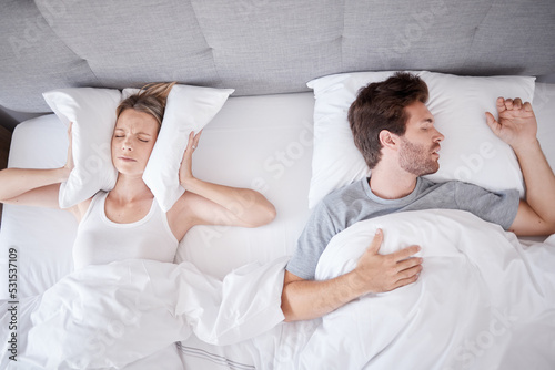Sleeping, snoring and wife with pillow on ears to stop noise from husband in bed with sleep problem. Insomnia, frustration and stress with tired woman in bedroom lying next to man with apnea photo