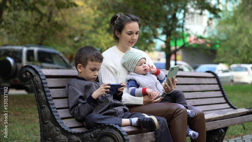Mom with two children are sitting on a bench in the park and are passionate about playing on the phone and watching cartoons