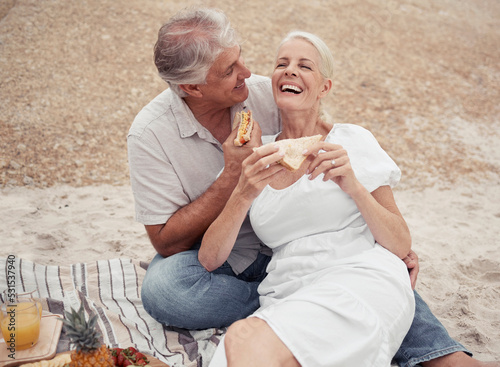 Love and healthy senior couple on picnic at a beach eating sandwich with funny conversation for wellness, healthcare or retirement. Outdoor elderly woman and man with cheese bread together on ground