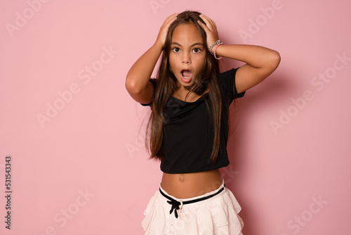 Fun portrait of cute brunette kid girl with hands on head.Youngster with open mouth and shocking facial expression isolated on pink background.