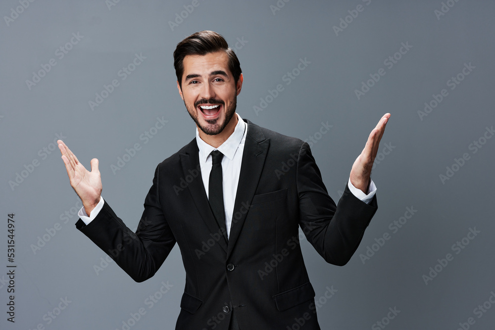 The man business in the suit is surprised and smiling a smile with his teeth raising his hands up against the gray background The man business in the suit is surprised and smiling a smile with his teeth raising his hands up against the gray background