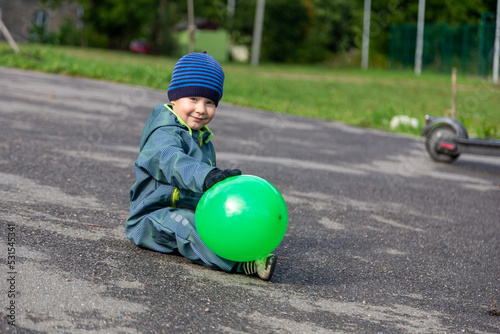 A two-year-old little toddler boy sits on a wet asphalt road with a green balloon in his hands in autumn on the street