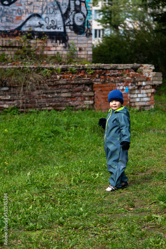 
A two-year-old little toddler boy stands on the grass near a ruined house. Childhood and destruction.

