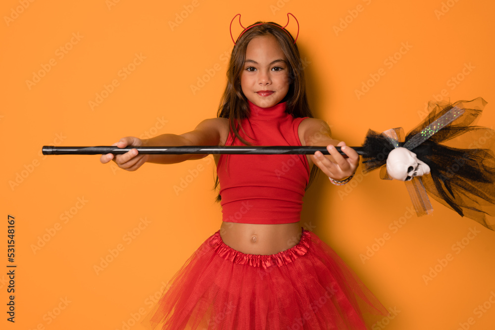 Beautiful cute little kid girl in red costume with devil´s horns holding a witch´s broom standing isolated over orange background. Halloween concept. Beautiful cute little kid girl in red costume with devil´s horns holding a witch´s broom standing isolated over orange background. Halloween concept.