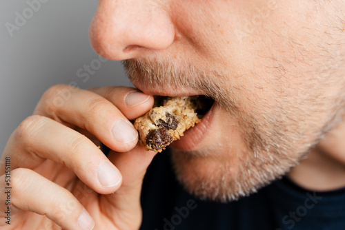 Close-up of middle-aged man with beard taking bite off healthy oatmeal raisin cookie, traditional and popular biscuit in America made of sugar, flour, rolled oats, eggs and dried raisins