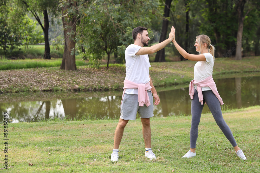 image of young caucasian couple man and woman enjoying in exercising and give me five outdoor with smiling faces and eyes contact together with green trees during summer time in the park