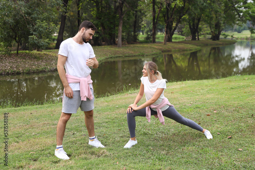 image of young caucasian couple man and woman enjoying in exercising and stretching outdoor with smiling faces and eyes contact together with green trees during summer time in the park