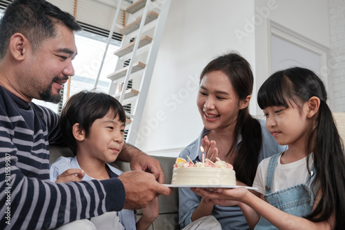 Happy Asian Thai family, young son is surprised with birthday cake, blows out candle, and celebrates joy party with parents and sister in living room together, wellbeing domestic home special event.