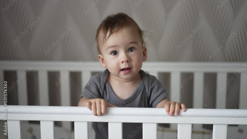 Beautiful curious little child rising on his feet in the white crib. Adorable baby looking into camera showing tongue. Beautiful curious little child rising on his feet in the white crib. Adorable baby looking into camera showing tongue.