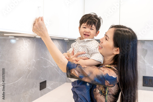 Female mother of east asian ethnicity with toddler son attempting to test cabinet door handles, signifying mom life and stay-at-home or working mom who is home decorating or being an interior designer