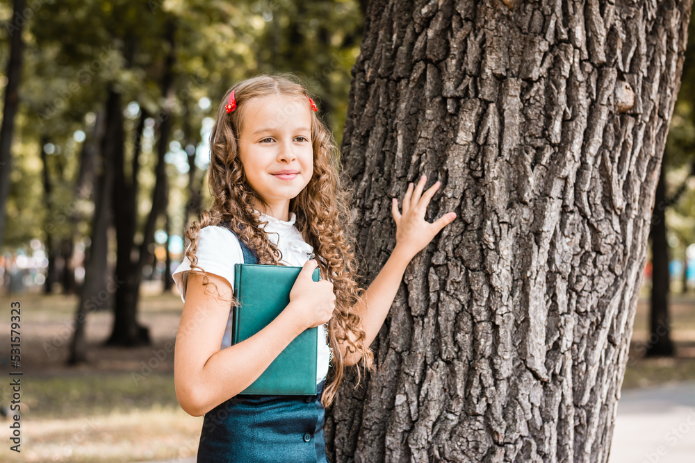 A cheerful female student in a school uniform with a book stands near a tree in the park on a warm day A cheerful female student in a school uniform with a book stands near a tree in the park on a warm day