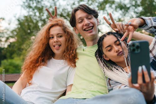 Three young smiling friends sitting on a park bench taking self-portrait on smartphones. Concept of friendship and technology. Self improvement and education